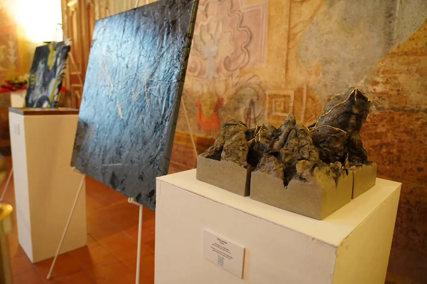 Sala dalle pareti affrescate con una tela scura e materica su un cavalletto e, accanto, su un plinto bianco, una scultura in ceramica a forma di catena montuosa percorsa da venature dorate
