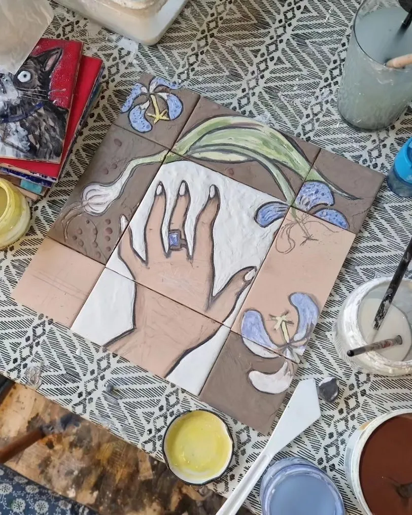 Lo stesso pannello di piastrelle dipinte disteso su un tavolo da atelier, tra barattoli di colore e pennelli
