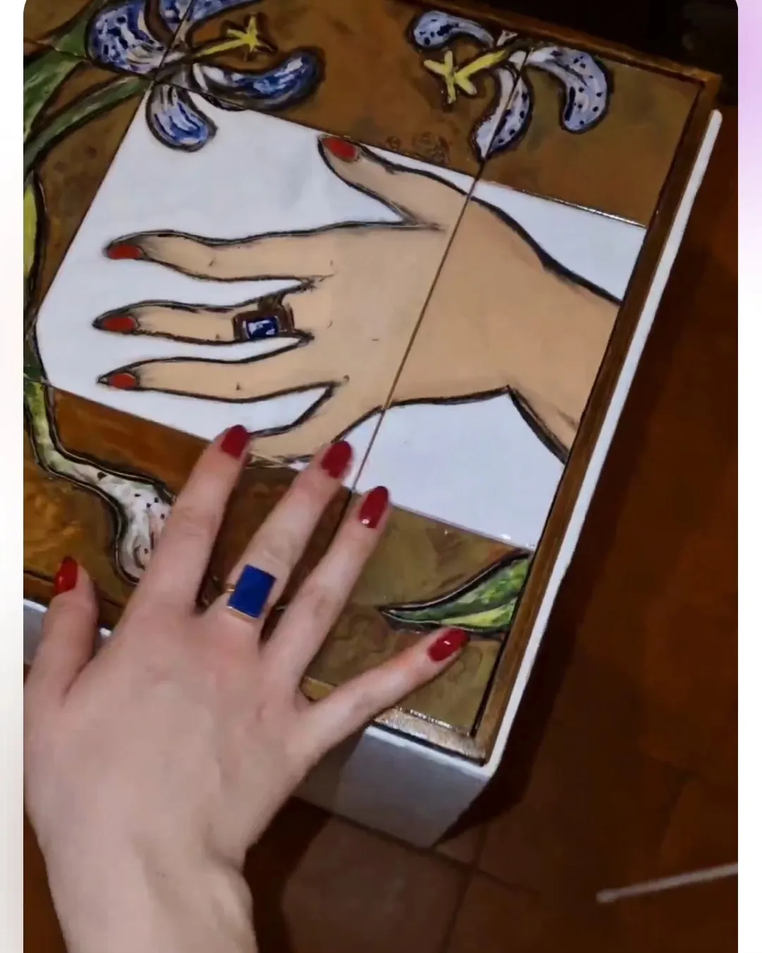 Una mano appoggiata su un pannello di piastrelle dipinte a mano con il disegno di una mano e di un giglio azzurro; al dito un anello con pietra blu