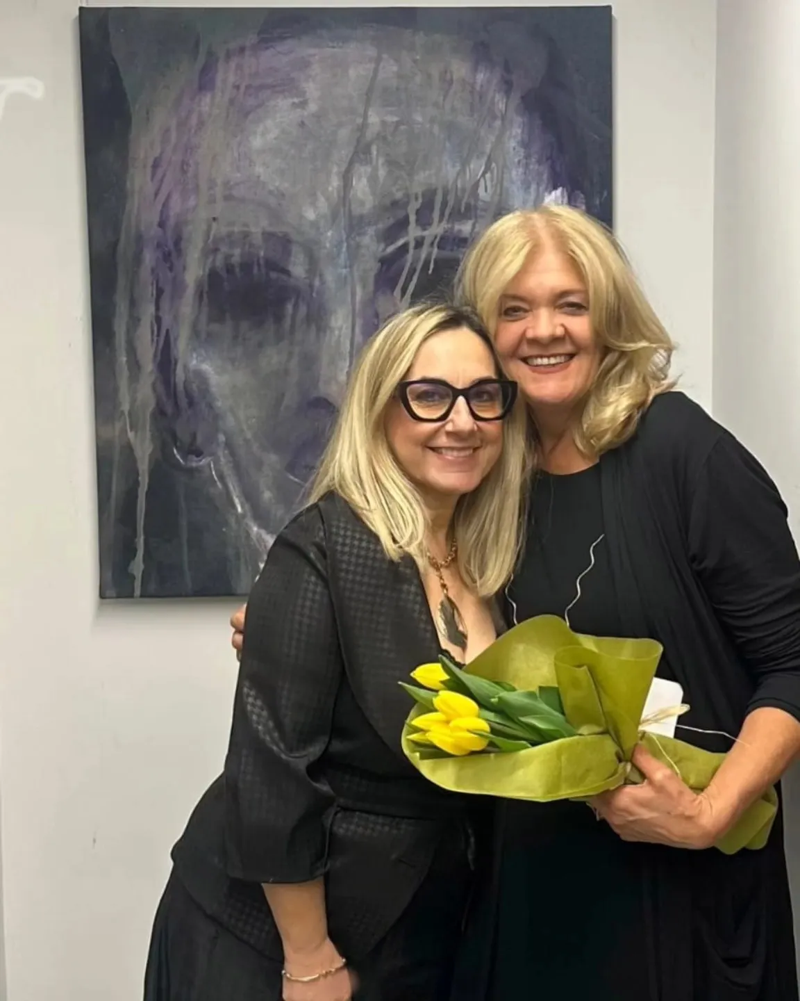 Due donne sorridenti e abbracciate al vernissage davanti a un ritratto scuro dai colori colati; una tiene un mazzo di tulipani gialli