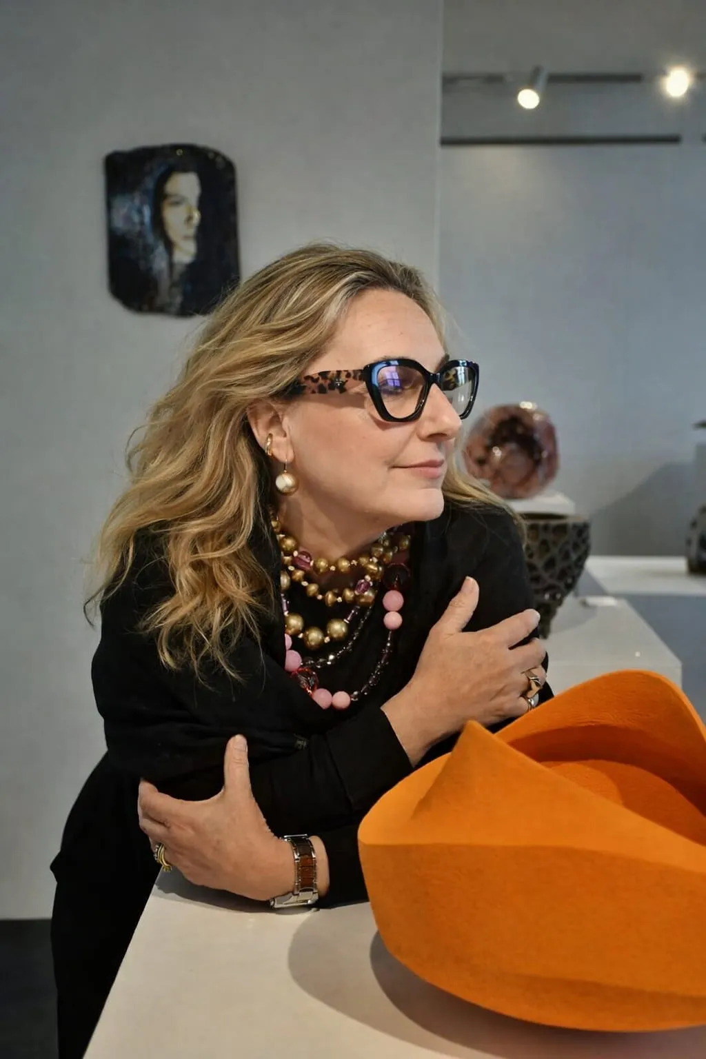 Rita Raimondi appoggiata a un basamento in una galleria, accanto a una scultura arancione; sullo sfondo altre opere e un ritratto alla parete